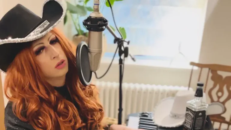 Amiss Tori drag queen sitting in front of a piana och microphone. Cowboy hat - red hair.