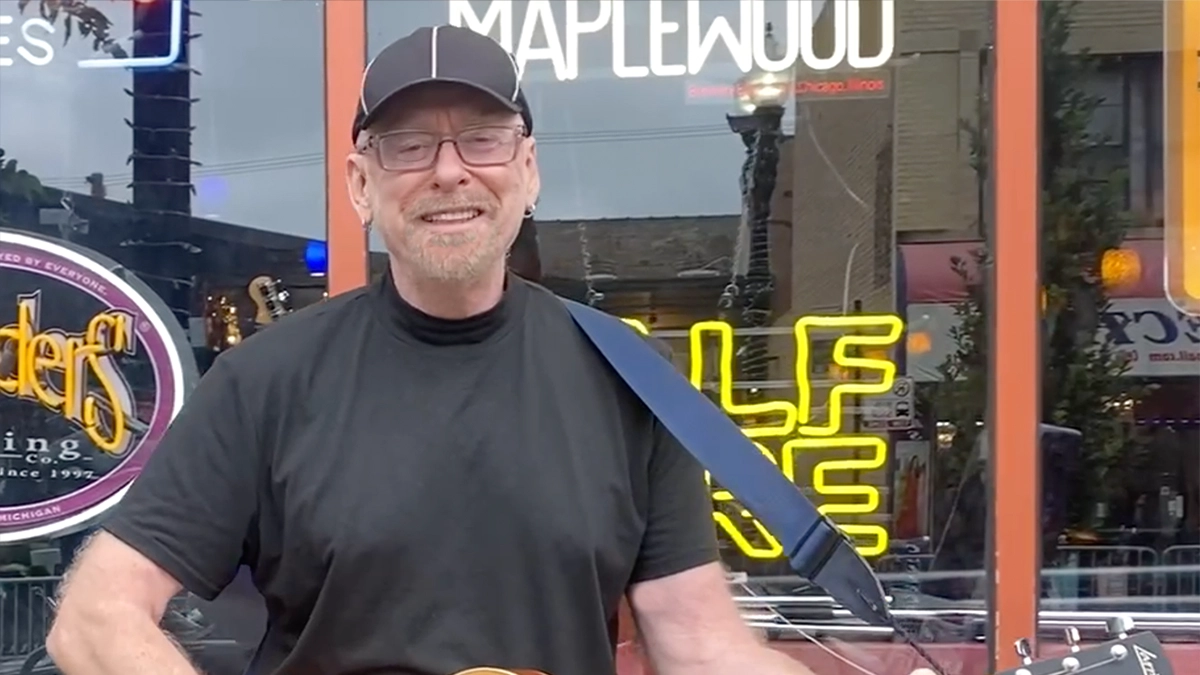 Scott Free is wearing a black t-shirt and black cap while playing the guitar. He has a grey beard and is also wearing metal-framed eyeglasses.