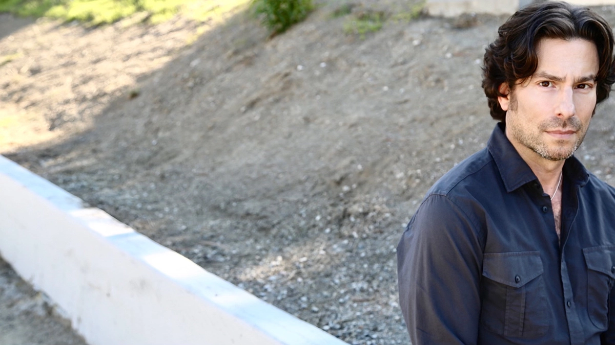 Picture of Jason Gould with garden trees in the background. Jason has dark brown eyes and dark curly hair. 3 days unshaved.