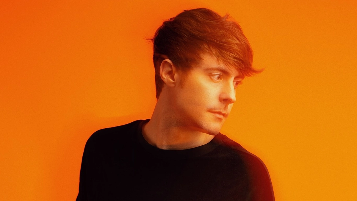 DallasK is standing, looking to the right, with an orange background, wearing a black sweatshirt and sporting brown hair.