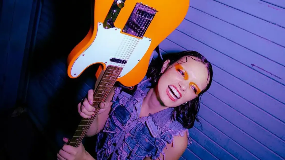 Ella Galvin holding a guitar upside down, against a purple background, wearing a jeans jacket.