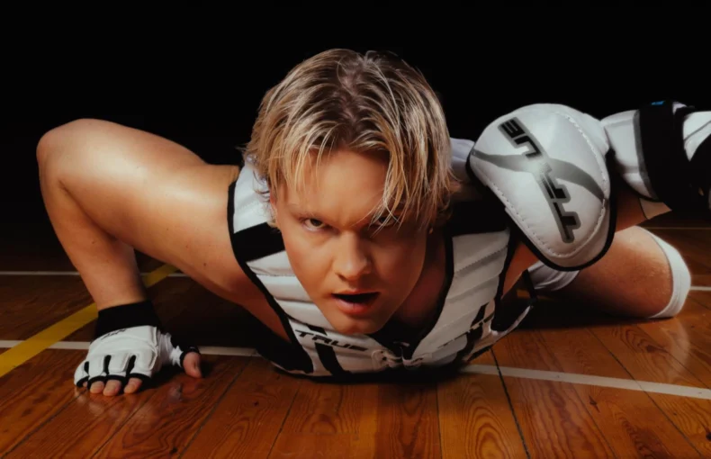 Benjamin in a dynamic pose, lying on the floor at the school gymnasium with his legs bent and head raised. The Benjamin has short blonde hair and wears a white and black sleeveless top with a thick black stripe around the armholes, paired with white and black shorts featuring a similar stripe at the waist. On his hands, he wear white fingerless gloves with black trim and ventilation holes at the fingertips and thumbs, secured by a strap that wraps over their forearm. Benjamin - Badabim
