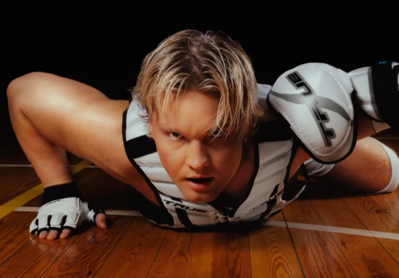 Benjamin in a dynamic pose, lying on the floor at the school gymnasium with his legs bent and head raised. The Benjamin has short blonde hair and wears a white and black sleeveless top with a thick black stripe around the armholes, paired with white and black shorts featuring a similar stripe at the waist. On his hands, he wear white fingerless gloves with black trim and ventilation holes at the fingertips and thumbs, secured by a strap that wraps over their forearm. Benjamin - Badabim