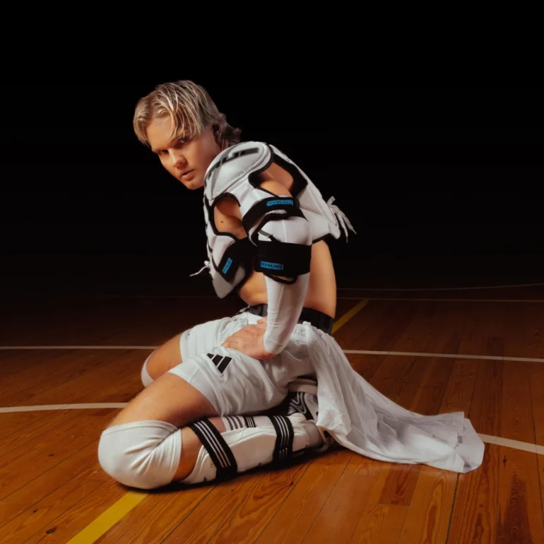The image depicts Benjamin in a sports uniform sitting on the floor of a gymnasium. He is wearing a white sports uniform with black and blue accents, consisting of a jersey, shorts, knee-high socks, and cleats. Shoulderpad has holes cut out around the neck, shoulders, and chest area, adding an edgy touch to the overall design. The shorts have "Adidas" black stripes on the front, The socks feature black stripes and a large "adidas" logo on the side, further emphasising the athletic theme. The cleats have thick black straps and white laces, giving the outfit a sporty and athletic feel. Benjamin's hair is styled in a messy, piecey look, with blonde highlights that complement the white and black color scheme of his uniform. In the background, the wooden floor of the gymnasium provides a warm and textured backdrop for the scene. The floor is made of light-colored wood planks, with white lines painted on them to create a basketball court or other athletic surface. Overall, the image presents a stylish and athletic Benjamin posing for a photo in a gymnasium. The sports uniform, with its combination of white, black, and blue accents, adds a touch of modernity and flair to the scene. The messy, piecey hair and bold fashion choices suggest that Benjamin is confident and expressive.